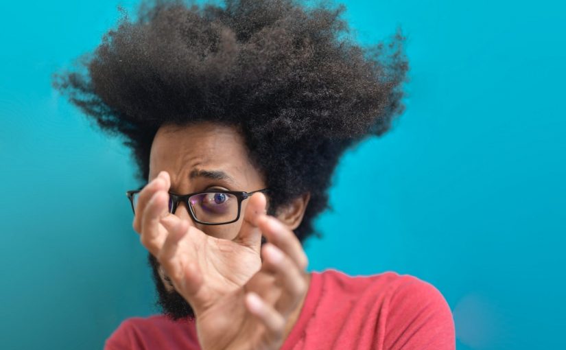 scared young ethnic man looking through hands on blue background in studio